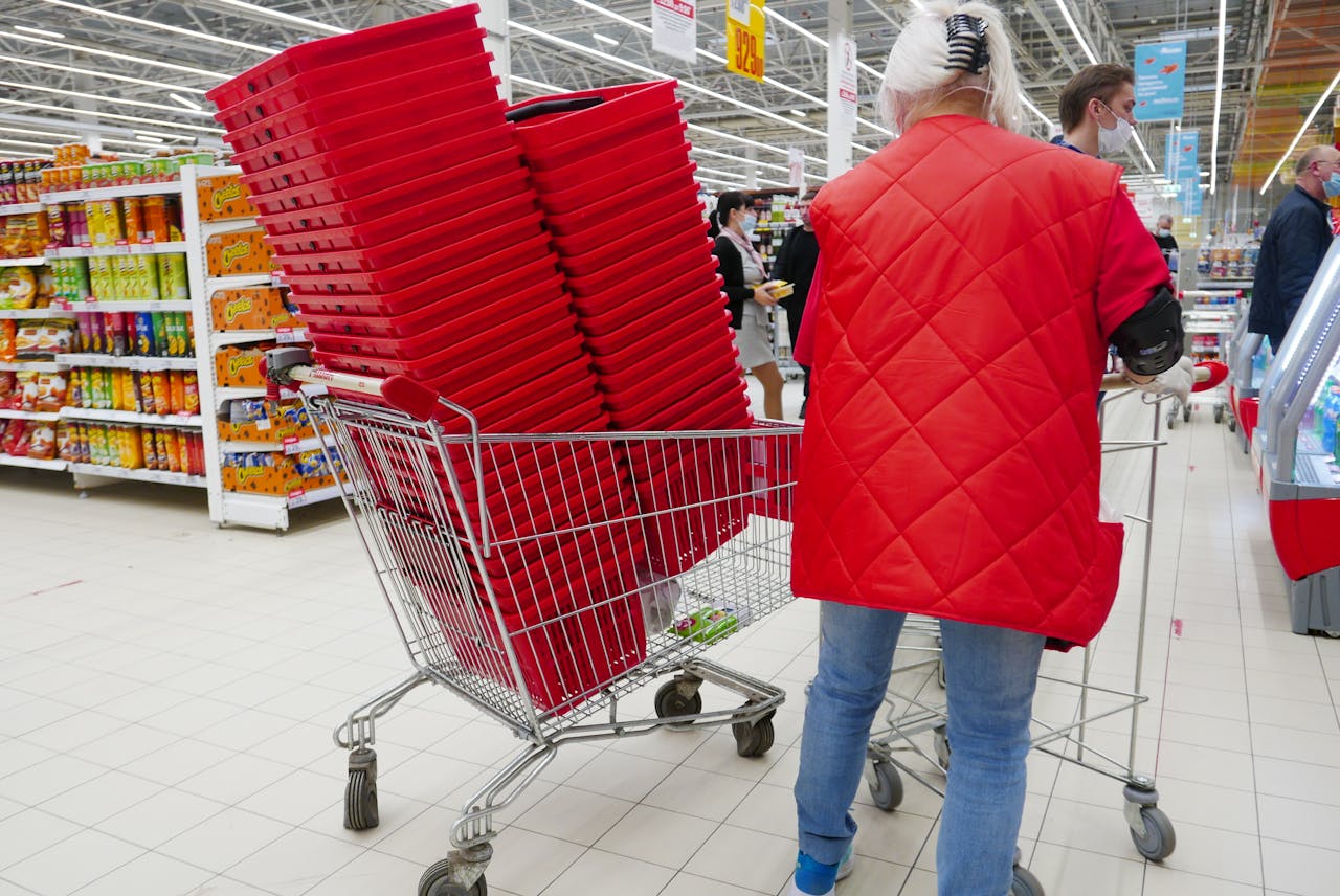 A shopper in a red coat with stacked red baskets inside a grocery cart in a supermarket.